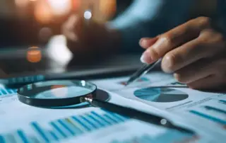 Eine Hand zeigt mit einem Stift auf ein Diagramm auf einem Dokument, das neben einer Lupe und einem Laptop liegt. Dies könnte auf die Vorbereitung oder Durchführung eines externen Audits hinweisen.